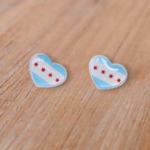 Op de afbeelding: Twee hartvormige oorbellen met een Chicago-vlagontwerp. Elke oorbel heeft een lichtblauwe en wit gestreepte achtergrond met vier rode sterren. De oorbellen liggen op een lichtbruin houten oppervlak.