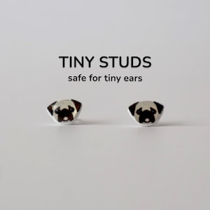 May include: Tiny stud earrings featuring a pug design. The earrings are silver-toned with a black and brown pug face. The text "TINY STUDS safe for tiny ears" is above the earrings.