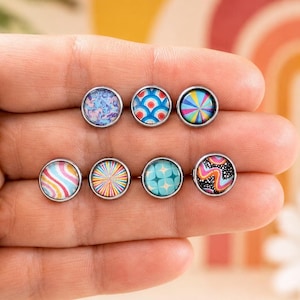 May include: Seven circular stud earrings with silver-toned metal settings. Each earring features a unique colorful graphic insert, including patterns of blue swirls, red and blue scales, multicolored sunbursts, wavy stripes, and black and pink waves.