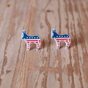 Peut inclure: Paire de boucles d'oreilles en forme d'âne avec un motif patriotique. Les boucles d'oreilles présentent un corps bleu avec des étoiles blanches et des rayures rouges et blanches, sur un fond en bois. Les boucles d'oreilles sont petites et détaillées.