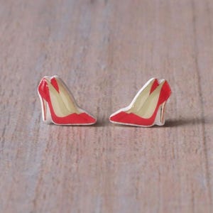 May include: A pair of miniature red high heel shoe earrings. The shoes have a white interior and a red exterior. The earrings are set against a light brown wooden surface.