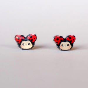 Puede incluir: Un par de pendientes de botón de mariquita. Cada pendiente presenta un diseño de mariquita roja con lunares negros y un contorno negro. Las mariquitas tienen una cara sonriente sobre un fondo blanco.