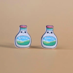 May include: Two cartoon-style earrings shaped like ranch dressing bottles. Each white bottle has a pink cap, a green and blue label with the word "RANCH", and a smiling face. The background is a solid tan color.