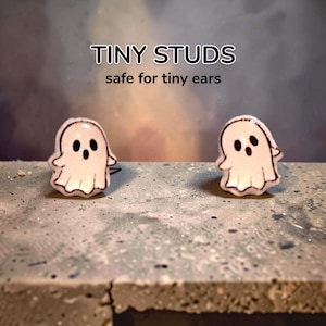 Może przedstawiać: Para kolczyków sztyftów w kształcie białych duszków z czarnymi oczami i ustami. Powyżej znajduje się napis TINY STUDS safe for tiny ears. Każdy duszek ma zaokrągloną głowę i drapowany korpus z wyraźnym czarnym konturem.