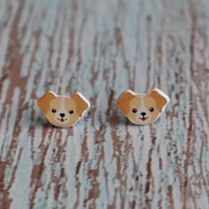 Op de afbeelding: Een paar houten oorstekers met een honden gezicht ontwerp. De oorbellen hebben een bruin en wit kleurenschema met zwarte ogen en een rode mond. De oorbellen zijn geplaatst tegen een lichtblauwe en bruine houten achtergrond.
