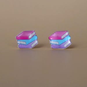 Peut inclure: Deux paires de boucles d'oreilles en forme de livre. Chaque boucle d'oreille présente une pile de trois livres aux tons rose, bleu et violet, avec des accents blancs. Les boucles d'oreilles ont une finition brillante et sont posées sur un fond neutre.