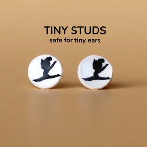 Puede incluir: Dos pendientes de botón blancos con la silueta negra de una gimnasta en espagat. Los pendientes son redondos y tienen un borde cosido. El texto "TINY STUDS safe for tiny ears" está encima.