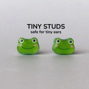 Könnte beinhalten: Zwei grüne Frosch-Ohrstecker mit rosa Wangen und weißen Augen. Der Text "TINY STUDS safe for tiny ears" steht über den Ohrringen. Auf weißem Hintergrund.