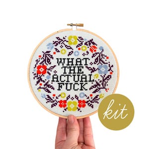 May include: A cross-stitch embroidery hoop with the phrase "WHAT THE ACTUAL FUCK" in black letters. The design includes a floral wreath with red, yellow, and blue accents. A gold circle with the word "kit" is in the lower right.