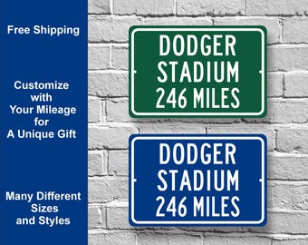 May include: Two rectangular signs with white text on a brick wall. The signs read "DODGER STADIUM 246 MILES". One sign is green, the other is blue. Text on the left side says "Customize with Your Mileage for A Unique Gift".