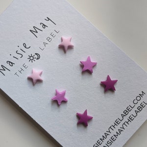 May include: A white card with the text "Maisie May THE LABEL" and six star-shaped objects in shades of pink. The stars are arranged on the card, with the brand's website and social media handle printed at the bottom.