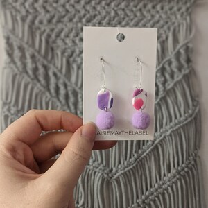 Puede incluir: Un par de pendientes colgantes con diseños abstractos de espiral en morado y rosa y un pompón lavanda en cada pendiente. Los pendientes se muestran en una tarjeta blanca con el texto "JaisieMayTheLabel".