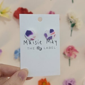May include: A pair of purple and white hexagonal earrings with a marbled pattern. The earrings are displayed on a white card with the text "Maisie May The Label".