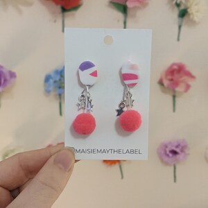 Può includere: Un paio di orecchini pendenti con motivo rosa e bianco, con pon pon rosa e ciondoli a forma di stella argentati. Gli orecchini sono esposti su una carta bianca con il testo "maisiemaythelabel".