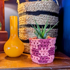 May include: A small green plant in a pink crocheted pot sits on a wooden table next to a yellow vase and a woven basket with black stripes.