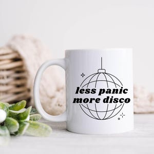May include: White ceramic coffee mug with a black outline of a disco ball and the text "less panic more disco".