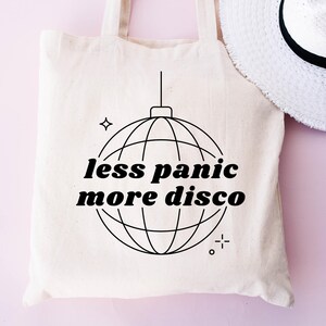 May include: A white tote bag with black text that reads "less panic more disco" and a black line drawing of a disco ball.