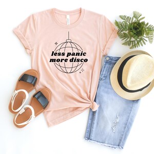 May include: A pink t-shirt with the text "less panic more disco" and a disco ball graphic. The shirt is paired with denim shorts, a straw hat, and sandals.