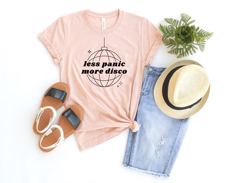 May include: A pink t-shirt with the text "less panic more disco" and a disco ball graphic. The shirt is paired with denim shorts, a straw hat, and sandals.