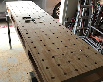Op de afbeelding: Een houten werkbank met een raster van gaten om gereedschap en klemmen aan te bevestigen. De werkbank is gemaakt van multiplex en heeft een natuurlijke houtkleur. De werkbank staat op twee bokjes.
