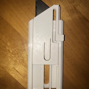 Op de afbeelding: Een witte cutter met een zwart mes. Het mes heeft een rechthoekige behuizing met een schuifmechanisme en een mes dat van bovenaf uitsteekt. Het mes is ontworpen voor het snijden van verschillende materialen.