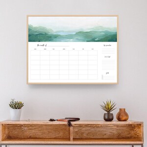 May include: A framed monthly calendar featuring a watercolour landscape design in shades of green and blue. The calendar has a simple grid layout with days of the week labelled at the top. The text "The month of" is at the top of the calendar. The calendar is hanging on a white wall above a wooden console table with a white wicker basket on the bottom shelf.