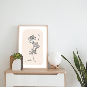 May include: A framed black and white line drawing of a marigold flower on a light pink background. The drawing is on a white dresser with a small potted plant and a white moon lamp.