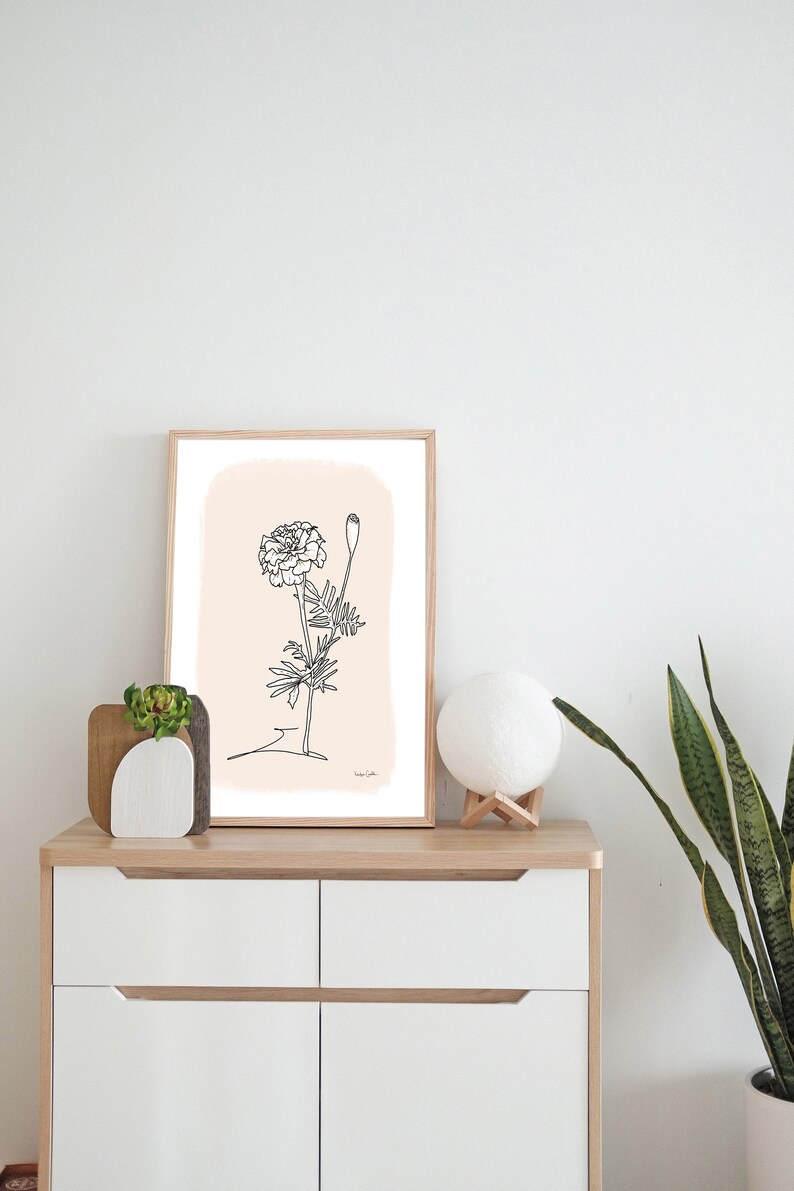 May include: A framed black and white line drawing of a marigold flower on a light pink background. The drawing is on a white dresser with a small potted plant and a white moon lamp.