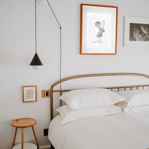 May include: A white bed with a gold metal headboard and a yellow and black patterned throw blanket. The bed is made with white linens and has two white pillows. There is a black lamp hanging from the ceiling and a small wooden side table with a white shelf.