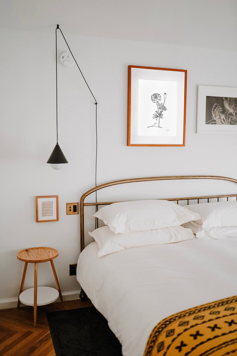 May include: A white bed with a gold metal headboard and a yellow and black patterned throw blanket. The bed is made with white linens and has two white pillows. There is a black lamp hanging from the ceiling and a small wooden side table with a white shelf.