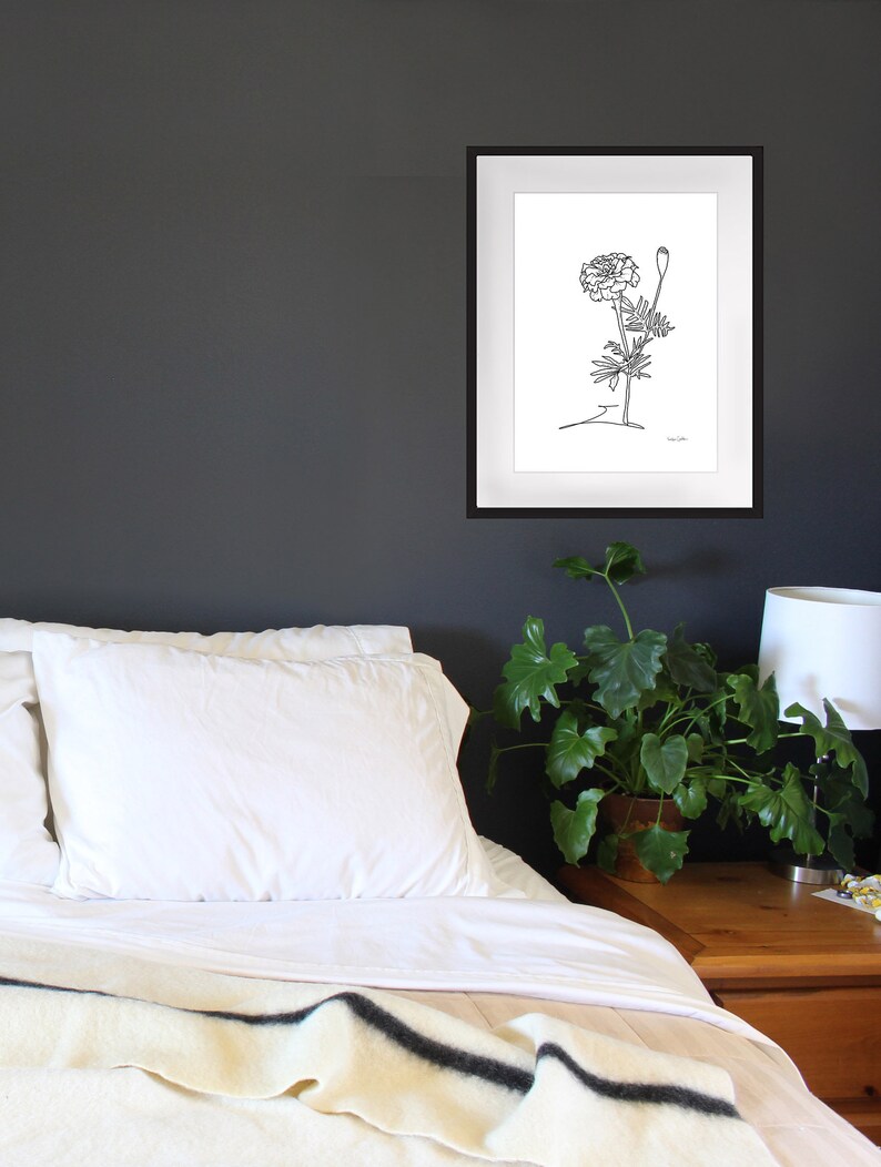 May include: A black and white line drawing of a marigold flower in a simple white frame. The frame is hanging on a dark gray wall above a white bed with a white blanket and a white pillow.