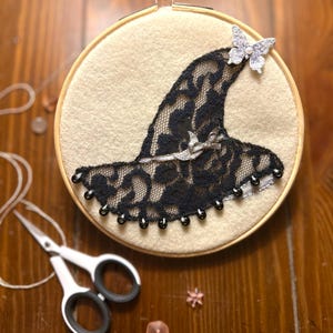 May include: An embroidery hoop featuring a witch hat design. The hat is crafted from black lace and adorned with a silver butterfly and beads. The background is a cream-colored fabric, and the hoop is made of wood. Scissors and beads are also visible.