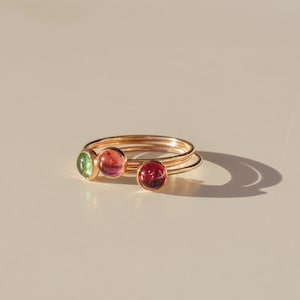May include: Three gold rings with different colored gemstones. The rings are stacked on top of each other. The gemstones are green, pink, and red.