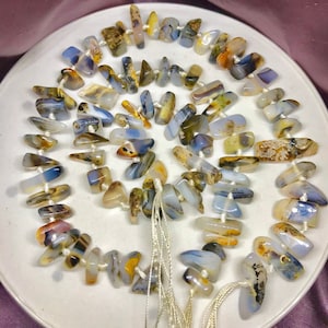 May include: A strand of blue, brown, and white agate beads with a natural, irregular shape. The beads are strung on white thread and are arranged in a circular pattern.