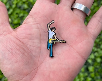 May include: A black enamel pin of a cartoon person playing a yellow electric guitar. The person is wearing a white shirt and blue jeans.