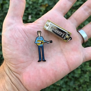 May include: A miniature Hohner harmonica and a guitar-playing figure pin are displayed. The harmonica is gold-toned with black text. The pin features a figure in a blue shirt and pants, playing a yellow guitar. The items are set against a blurred green background.