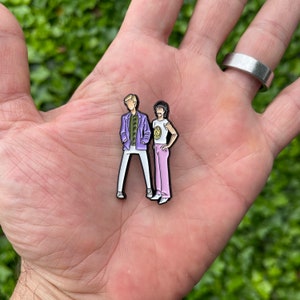 May include: Two enamel pins depicting cartoon figures of two people. The person on the left is wearing a purple jacket, a green shirt, white pants, and brown shoes. The person on the right is wearing a white shirt with a yellow circle design, pink pants, and brown shoes.