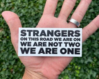 May include: White sticker with black text that reads "STRANGERS ON THIS ROAD WE ARE ON WE ARE NOT TWO WE ARE ONE".