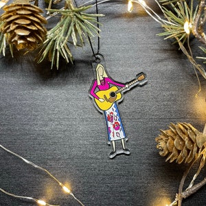 May include: A clear acrylic Christmas ornament featuring a cartoon illustration of a woman playing a yellow acoustic guitar. She is wearing a pink shirt and a colorful patterned skirt.