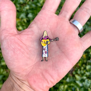 May include: A colorful enamel pin featuring a woman with blonde hair playing a yellow acoustic guitar. She is wearing a purple shirt and a white and blue patterned skirt.
