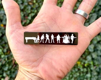 May include: A black metal bar with a white silhouette of a band playing music. The band includes a keyboard player, a guitarist, a bassist, a drummer, and a singer.
