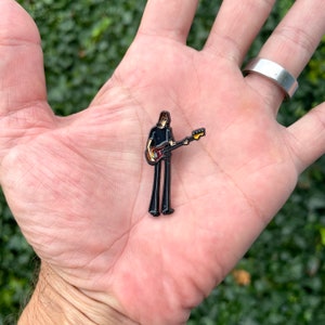 May include: Black and white enamel pin of a person playing a bass guitar. The person is wearing a black shirt and black pants. The bass guitar is red and yellow.