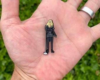 May include: Black enamel pin of a woman with blonde hair wearing a black dress and holding a microphone. The pin is on a hand.