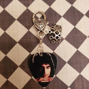 May include: A silver charm with a guitar pick featuring a photo of Freddie Mercury. The charm has a silver heart-shaped clasp and a silver crown.