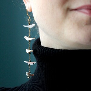 May include: Dangling earrings featuring white bird charms suspended from gold-colored chains. Each earring has multiple bird-shaped pendants, with small black beads and gold-colored accents. The earrings are a unique accessory.