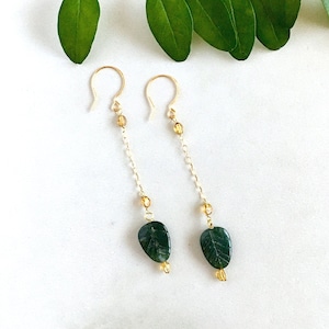 May include: A pair of gold-colored earrings with a leaf-shaped green stone pendant. The earrings have a delicate chain design with small yellow beads. The background is white with green leaves.