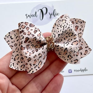 May include: A light pink and brown leopard print hair bow with a gold glitter center. The bow is held in a hand and sits on a white card with the text "sweet P jolie".