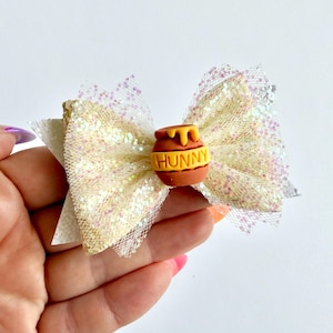 May include: A cream-coloured glitter hair bow with a decorative honey pot charm. The bow has a layered design with a mesh overlay and a small, brown honey pot with the word "HUNNY" in yellow.