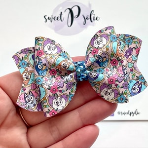 May include: A pink and blue patterned hair bow with a glitter center. The pattern features cartoon dogs and hearts. The bow is tied with a ruffle.