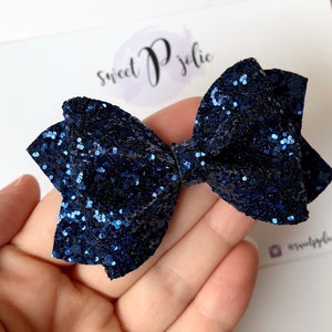 Könnte beinhalten: Eine marineblaue Glitzer-Schleife, ein Haaraccessoire, wird auf einer weißen Karte gezeigt. Die Schleife besteht aus einem glitzernden Material und hat eine klassische Schleifenform. Auf der Karte steht der Text "sweet P jolie".